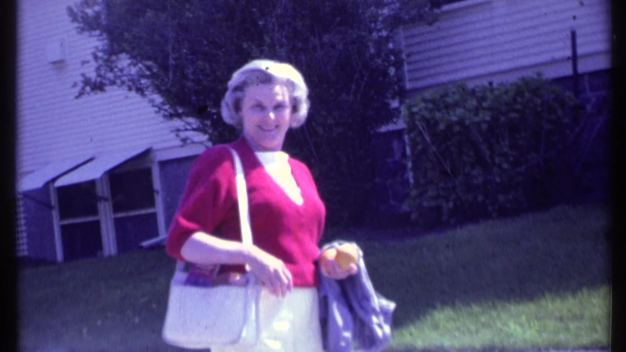 Adult woman in 1984 Vermont wearing a red sweater, white pants, and white purse looks toward the camera beside a house and lawn.
