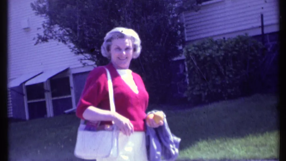 Adult woman in 1984 Vermont wearing a red sweater, white pants, and white purse looks toward the camera beside a house and lawn.