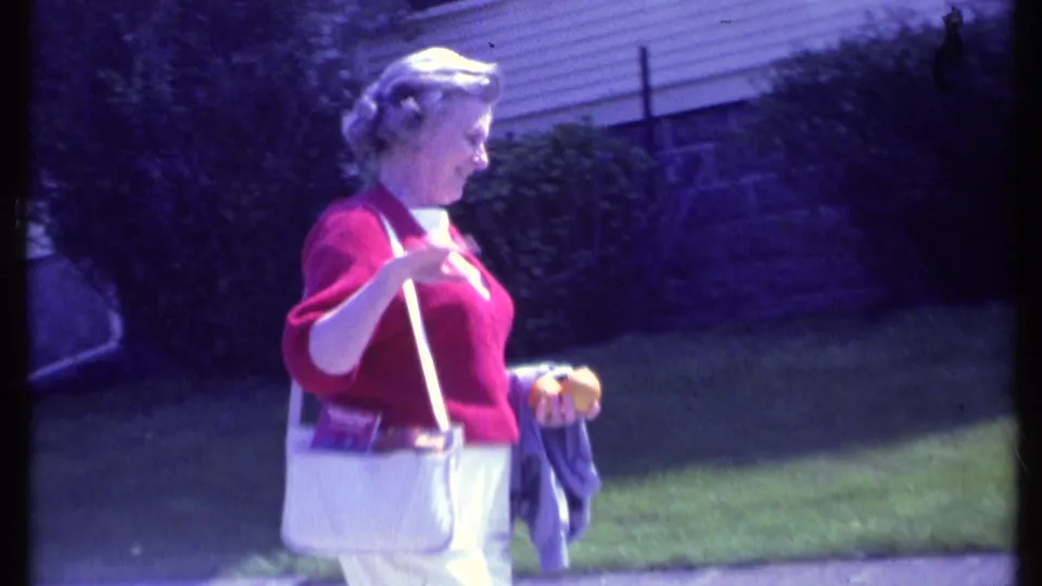 Adult woman in 1984 Vermont walks along a suburban sidewalk, raising her hand with a white purse over her shoulder.