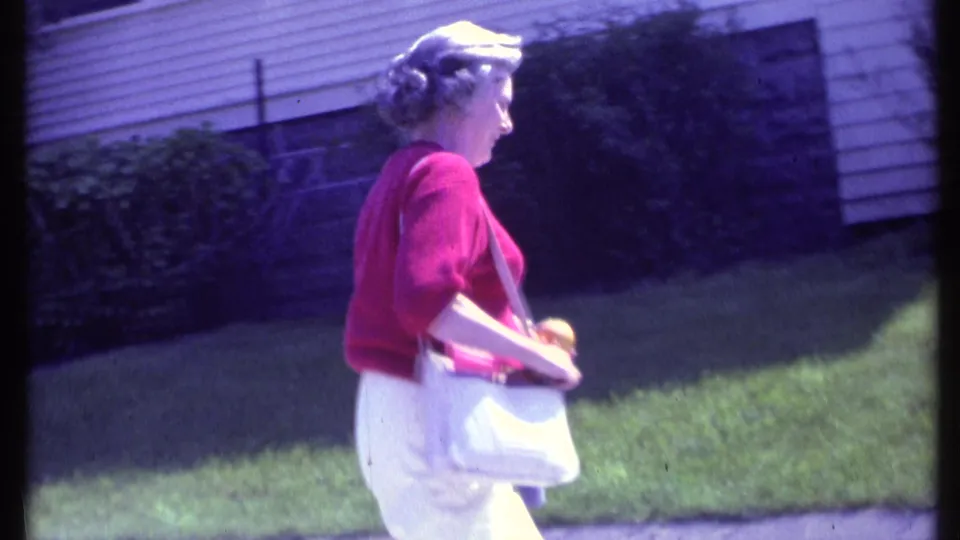Side view of the adult woman in a red sweater and white pants walking past a green lawn in 1984 Vermont home-movie footage.