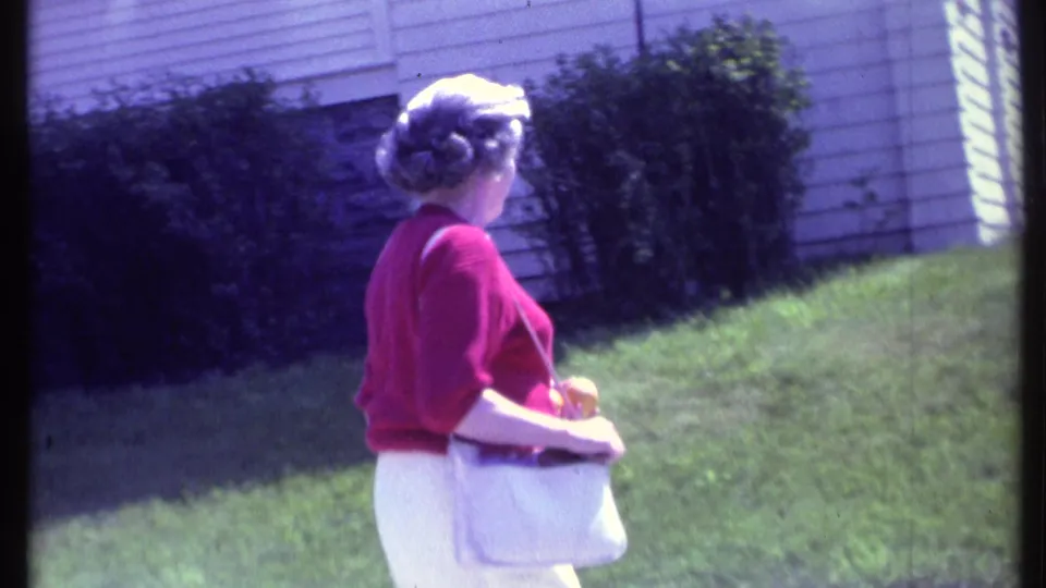 Adult woman in red and white is seen from behind as she walks away along a residential sidewalk in Vermont home-movie footage.