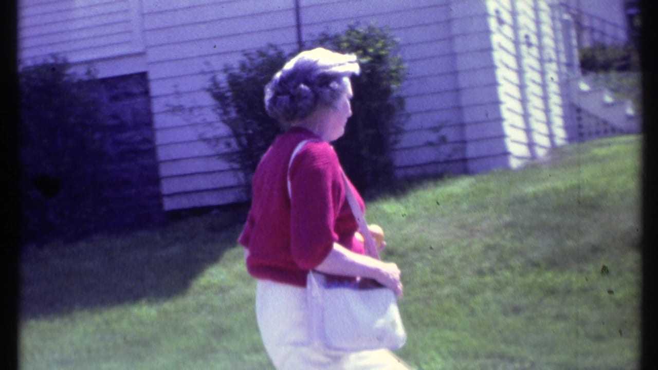 Adult woman in a red sweater, white pants, and white shoulder purse walks across a Vermont lawn in 1984 home-movie footage.