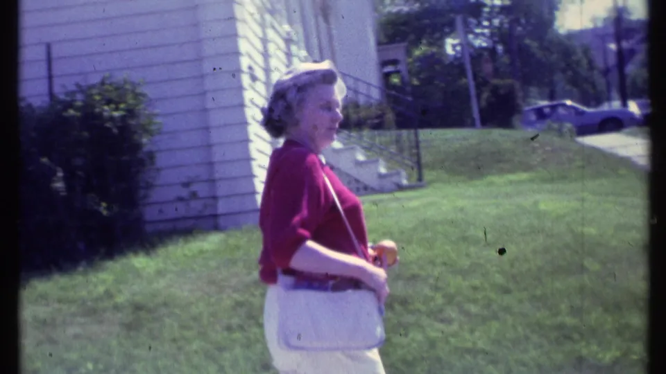 Adult woman in red and white clothing walks through a Vermont residential yard, carrying a white purse and jacket.