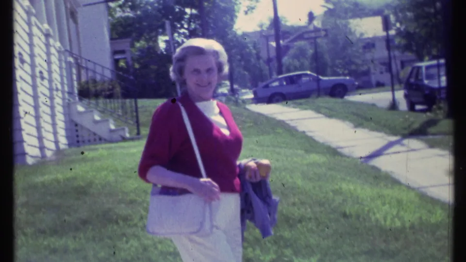 Adult woman smiles toward the camera while walking across a Vermont lawn in 1984 residential home-movie footage.