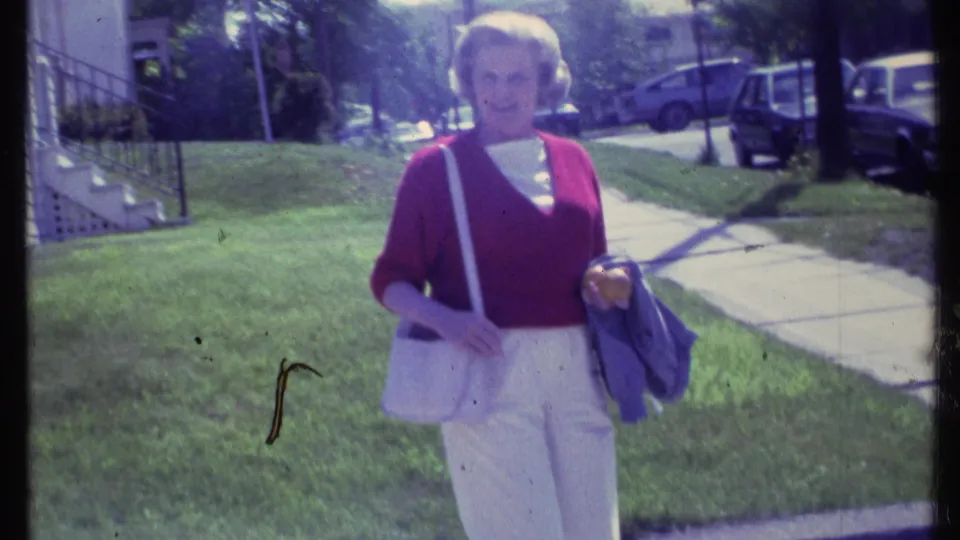 Adult woman in a red sweater and white pants approaches the camera in a Vermont residential yard, holding a jacket and purse.