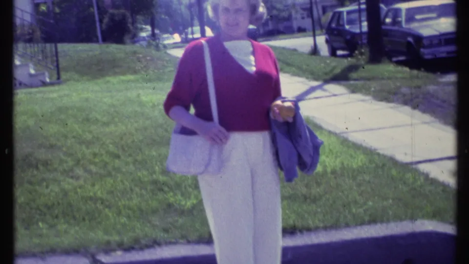 Adult woman in Vermont, 1984, wearing a red cardigan and white trousers, carrying a light purse and jacket on a residential sidewalk.