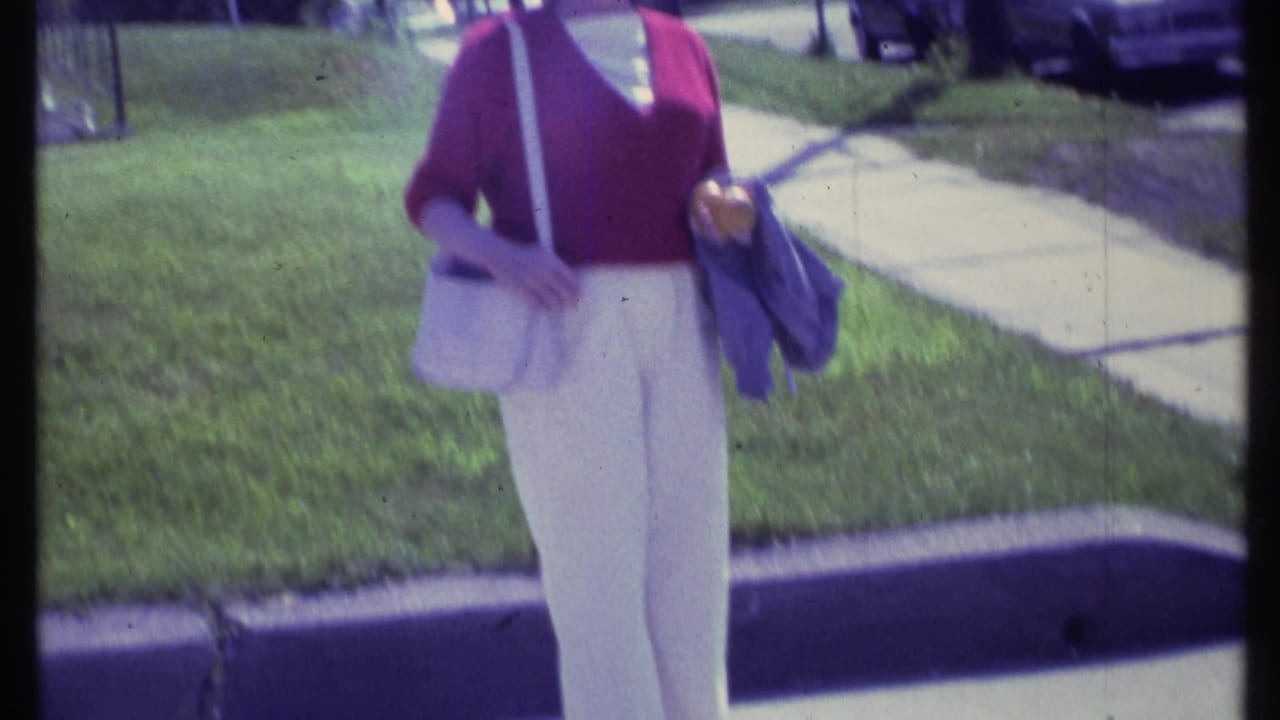 Closer view of the woman from torso down as she holds a purse and jacket beside a residential sidewalk.