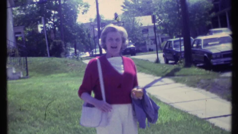 Adult woman in a red cardigan appears to look toward the camera on a Vermont residential sidewalk in 1984, with cars and homes behind her.