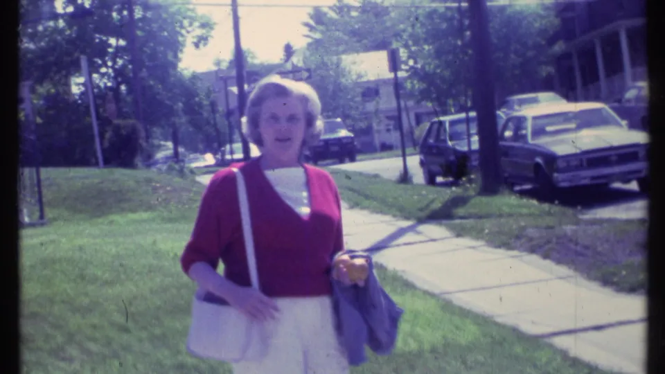 Vermont 1984 home-movie frame of a woman in a red cardigan and white trousers standing along a suburban sidewalk with parked cars nearby.