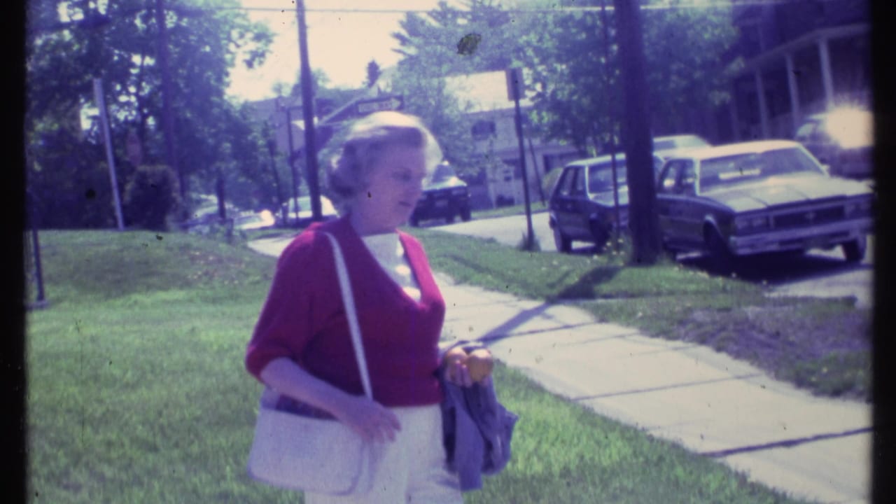 In supplied 1984 Vermont footage, a woman in a red cardigan walks beside lawns, parked cars, and houses.