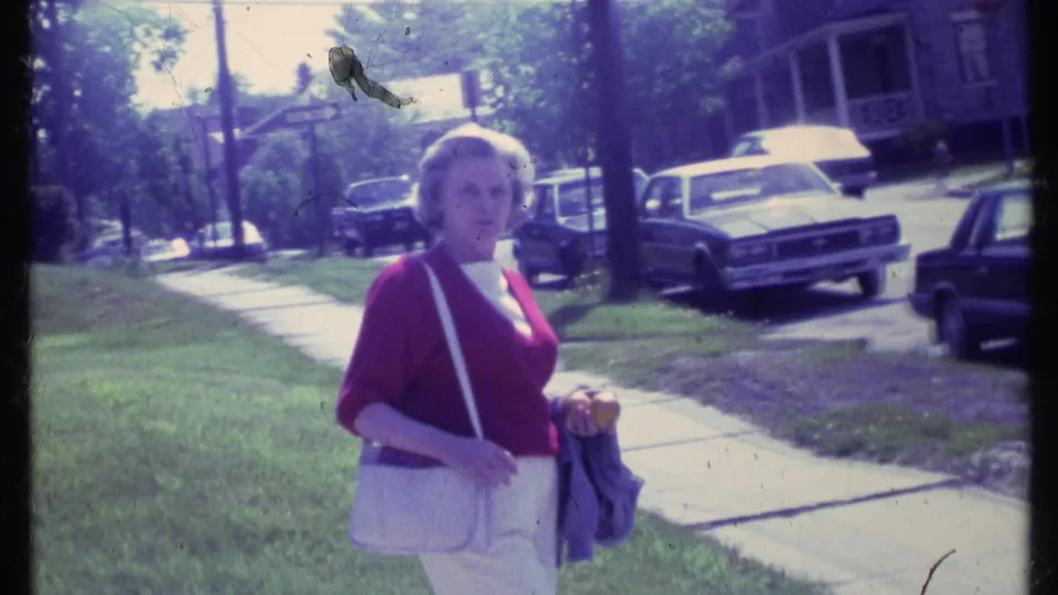 The woman looks toward the camera on a residential Vermont sidewalk, with cars and houses behind her.