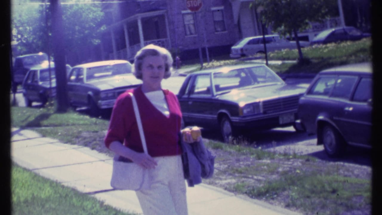 Adult woman in a red sweater and white pants walks on a Vermont sidewalk in 1984 with parked cars and brick homes behind her.