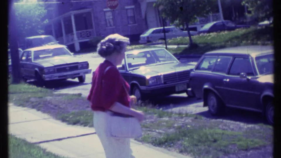 The woman continues along the sidewalk in profile, with a stop sign, lawn, parked cars, and brick houses visible in the background.