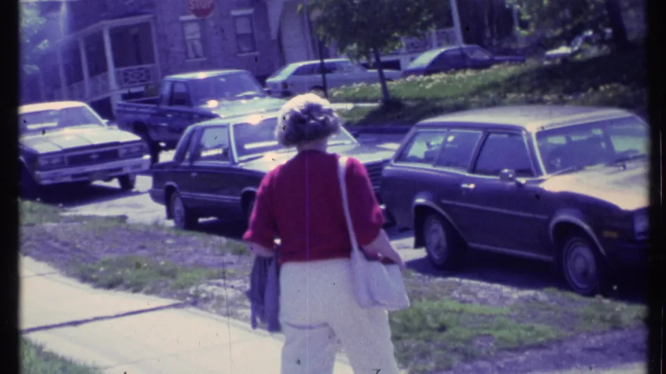 Seen from behind, the woman walks along a residential sidewalk with a dark station wagon, sedans, grass, and brick homes nearby.