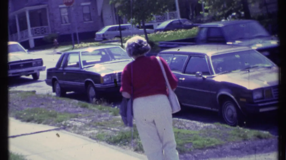 The woman walks farther down the Vermont sidewalk in 1984, passing a dark parked station wagon and other cars on a quiet residential block.