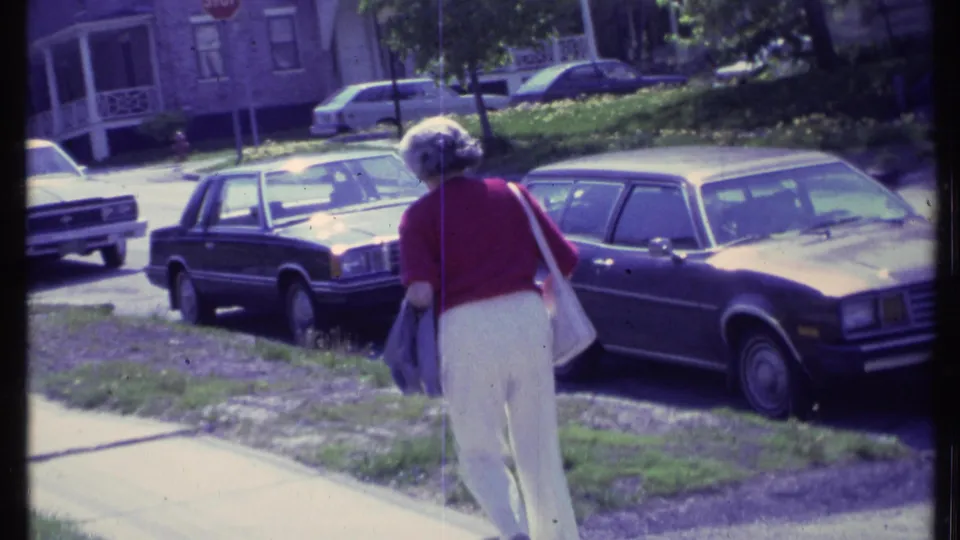 In Vermont 1984 home-movie footage, a woman with a purse walks away along a residential sidewalk past curbside cars.