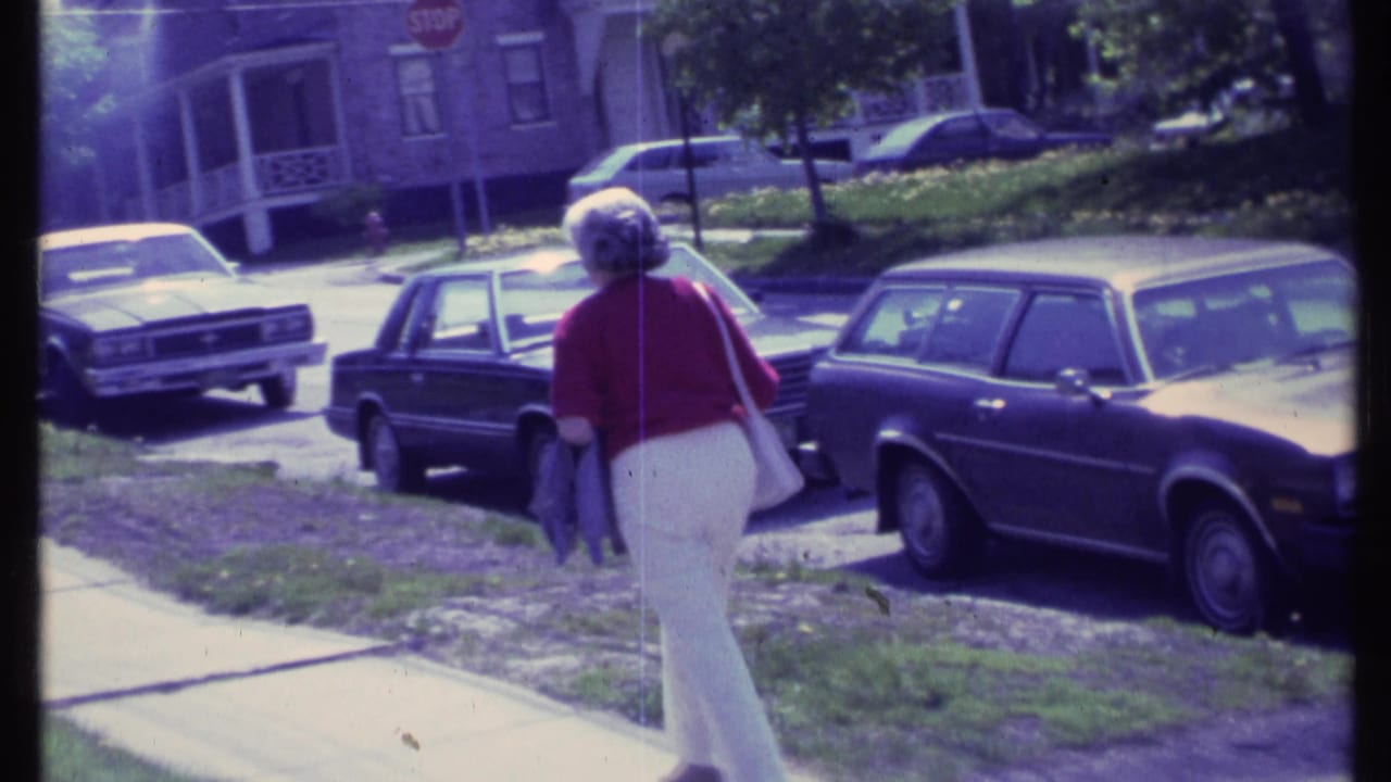 Woman continues down the sidewalk with parked cars and houses across the street.