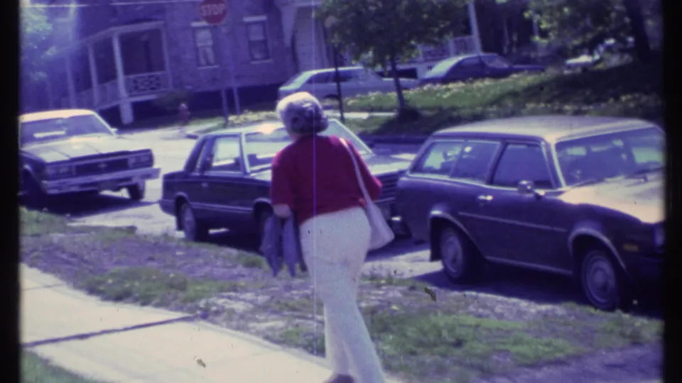A woman carrying a purse and jacket moves away on a Vermont residential sidewalk, with 1980s cars and houses nearby.