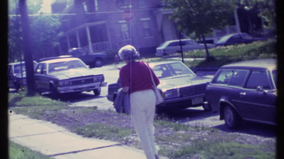 The 8mm frame shows the woman from behind on a sidewalk, with lawns, curbside vehicles, and house fronts in Vermont.