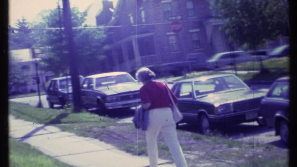 Near the end of the Vermont 1984 shot, the woman leans slightly with her purse and jacket as cars and houses remain in view.