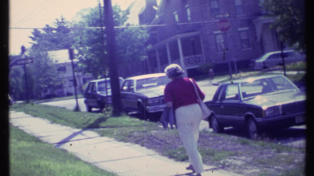 In supplied 1984 Vermont footage, an elderly woman carries a shoulder purse while walking past parked cars and houses on a sunny sidewalk.