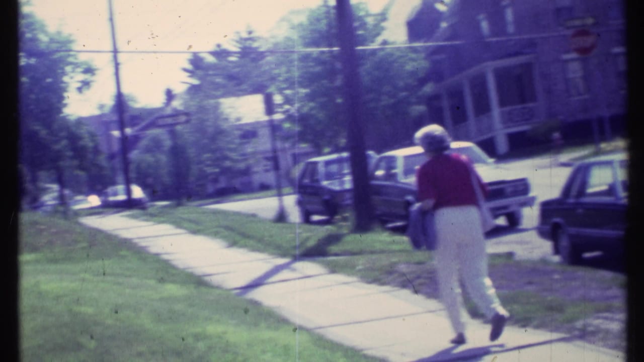 Woman continues down the sidewalk beside grass, trees, and parked cars.