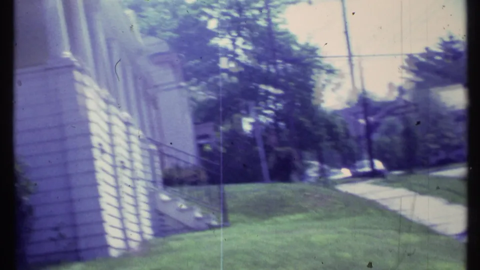 A handheld frame shifts toward a light-colored house wall and lawn, with the sidewalk and tree-lined street still visible.