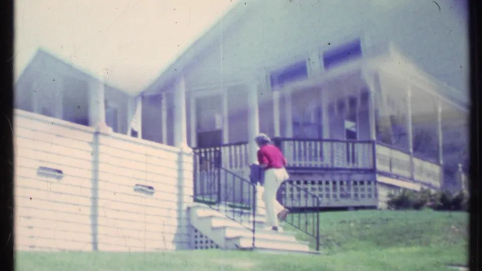 In Vermont 1984 home-movie footage, a woman in a red shirt and light pants climbs stairs toward a white house porch.
