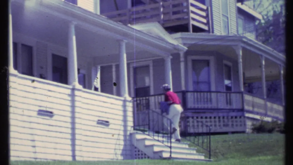 A woman in a red shirt ascends the stairs beside a dark railing, with a white Vermont house and porch behind her.