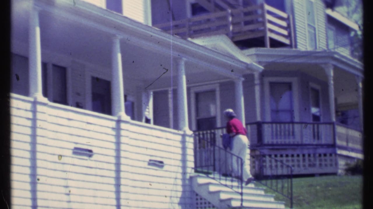 In 1984 Vermont home-movie footage, a woman in a red top approaches the front porch of a white clapboard house.