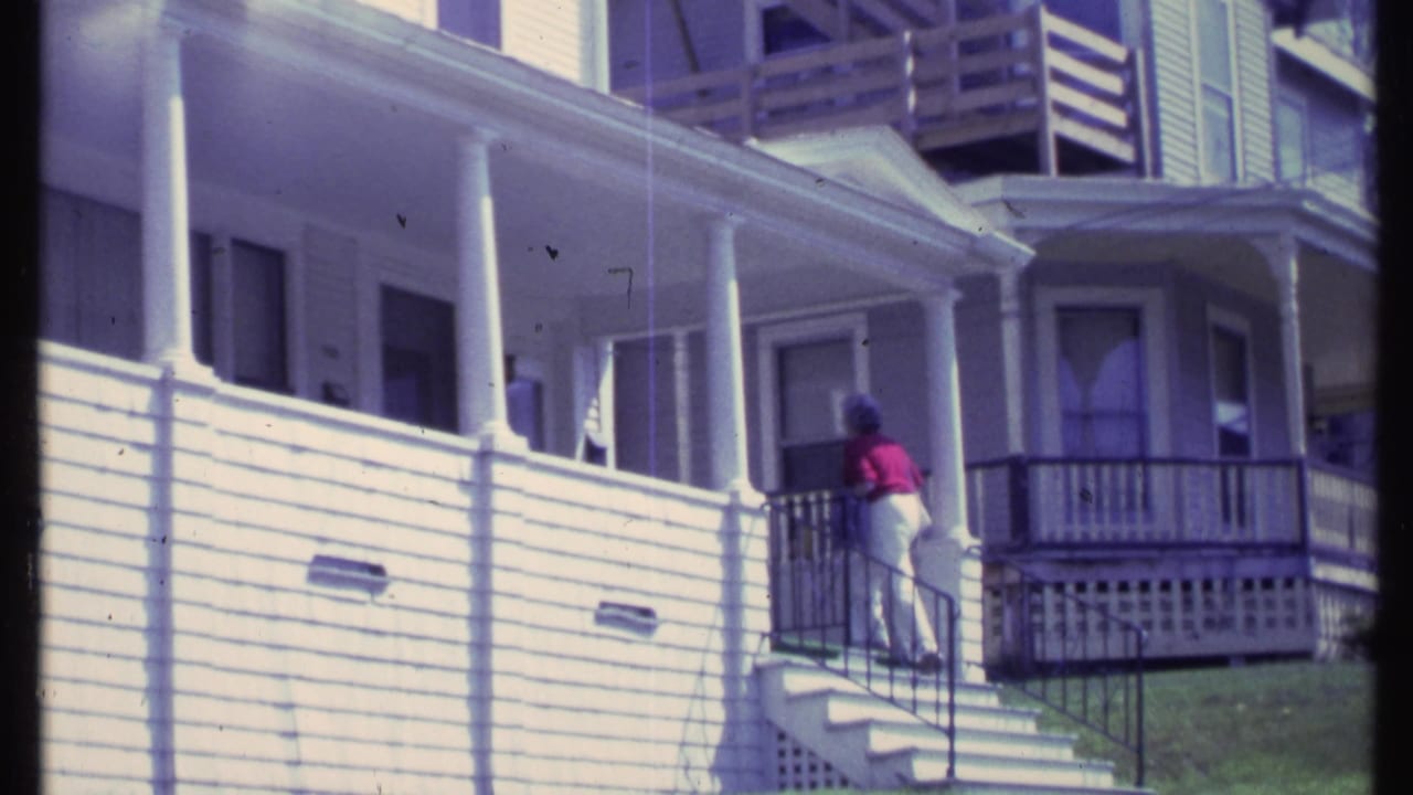 Woman stands near the top of the steps beside the porch railing of a white house.