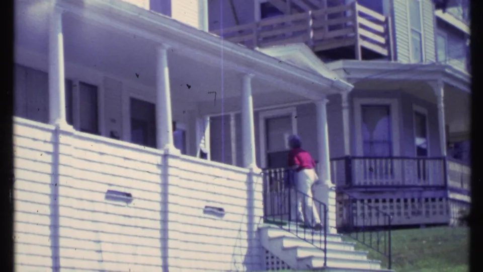 The woman reaches the upper steps by the porch railing on a white clapboard Vermont house in 1984 footage.