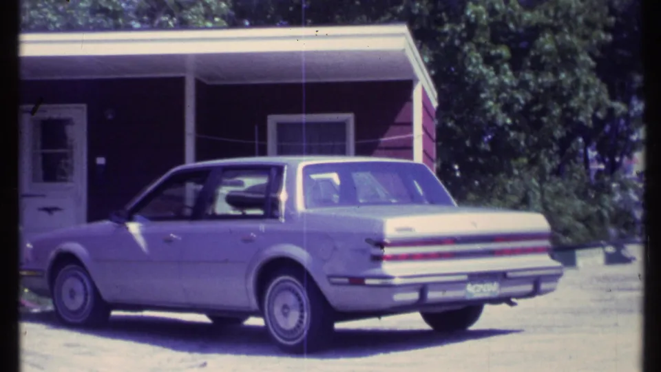 A light-colored 1980s sedan sits beside a Vermont house with a shaded porch and trees in the background.