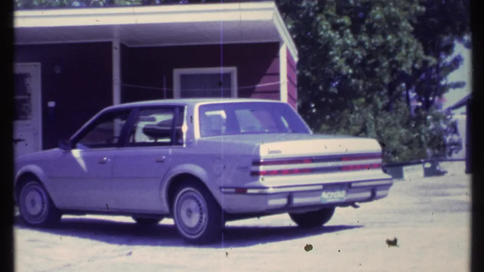 1984 Vermont home-movie frame showing a light-colored four-door sedan in a sunny residential driveway beside a red-sided house.