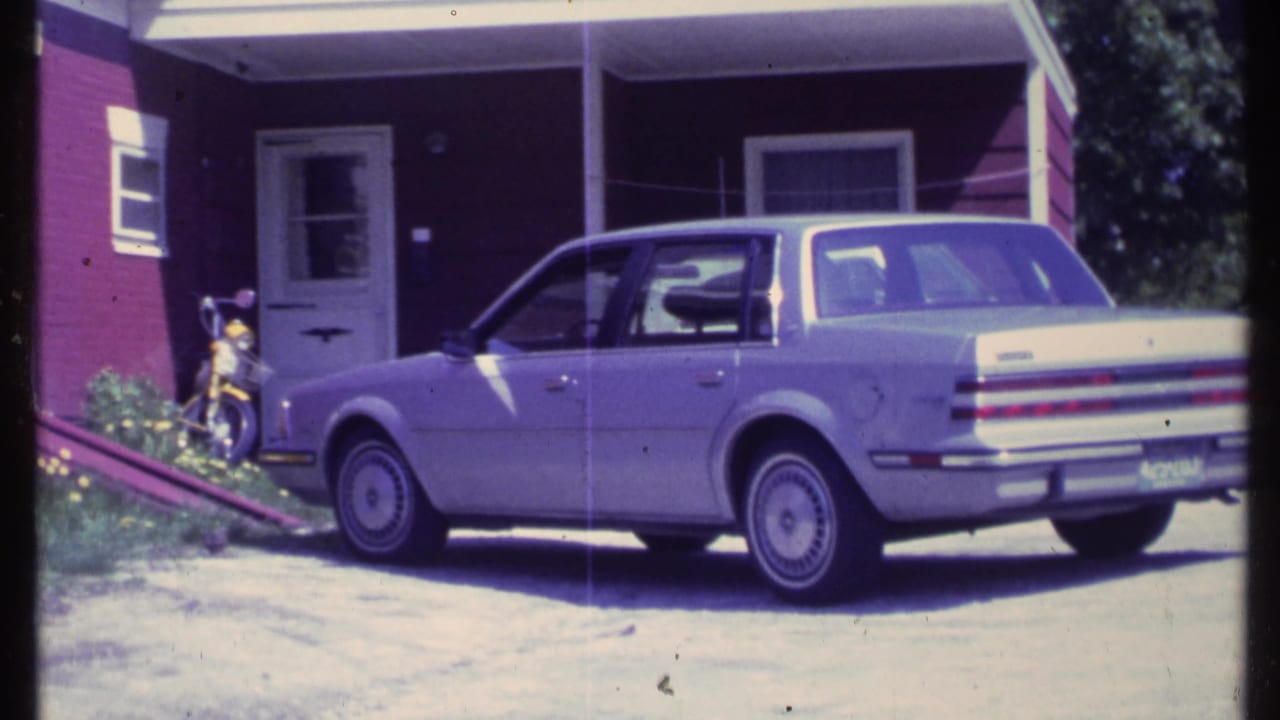 Sedan beside a red-sided home, with a small motorcycle or bicycle near the entry.