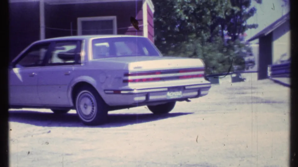 1984 Vermont frame focusing on the rear quarter of a light-colored sedan parked beside a red residential building.