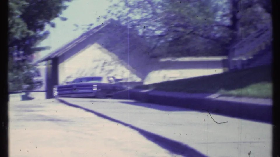1984 Vermont home-movie frame showing a driveway, neighboring garage, trees, and a partially visible parked vehicle in bright sun.