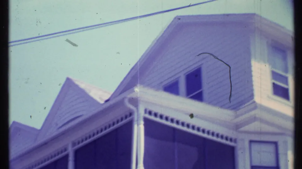 1984 Vermont home-movie frame showing the upper stories of a white house with porch roof, small windows, trim, and overhead wires.