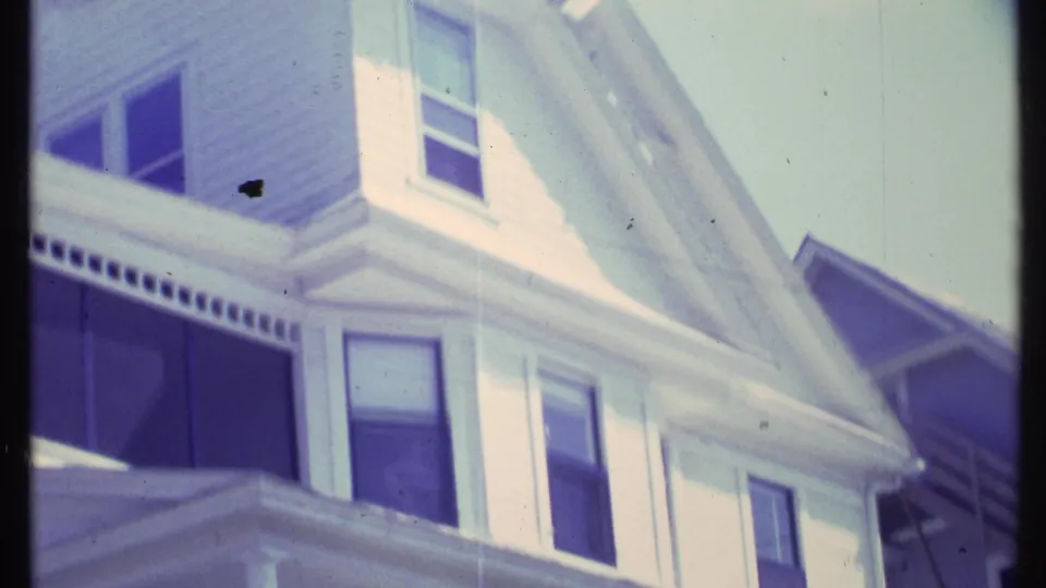 1984 Vermont home-movie frame of a white house facade, showing upper windows, a prominent gable, and dark porch opening.