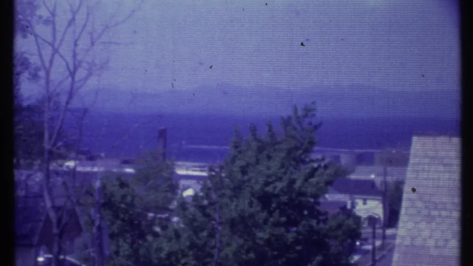 1984 Vermont home-movie frame showing distant mountains, tree silhouettes, rooftops, and a nearby shingled roof edge.