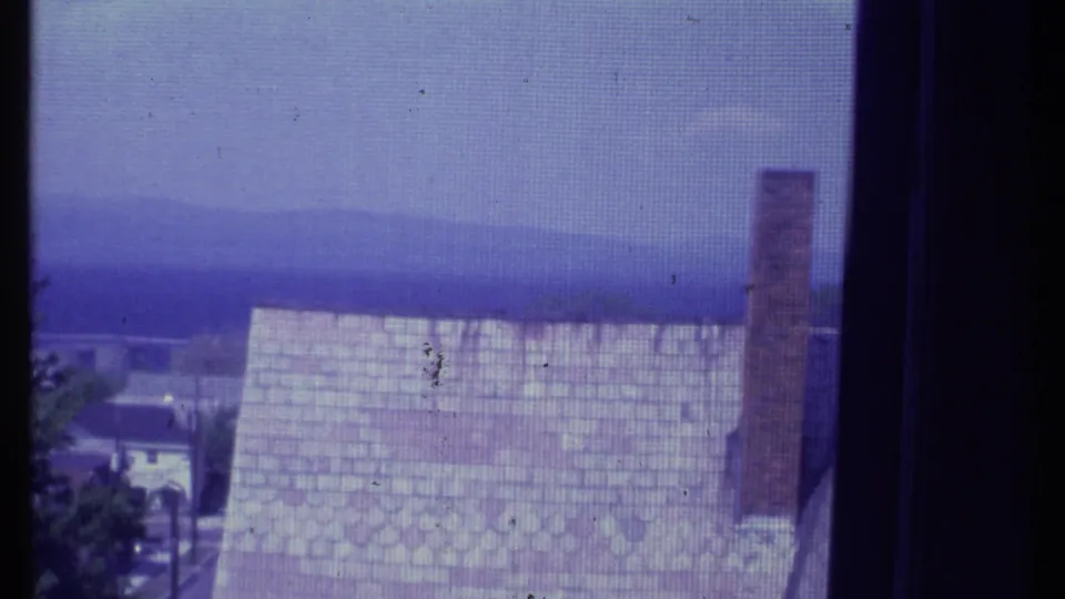 1984 Vermont 8mm frame showing a shingled roof, tall brick chimney, trees, and a distant mountain horizon through a screen.