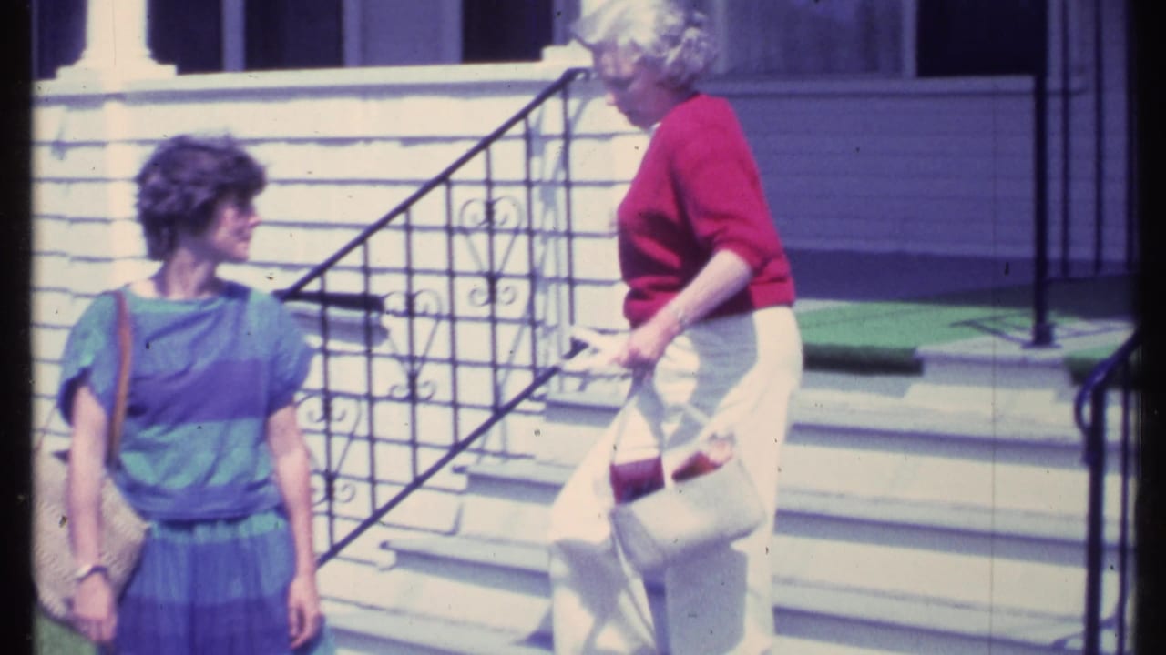 Two adult women stand near front steps outside a white clapboard house.