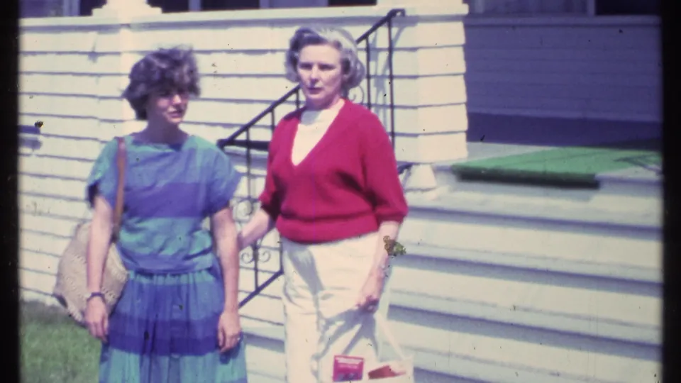 Two adult women in 1980s clothing stand near a white clapboard house in Vermont, with steps and railing behind them.
