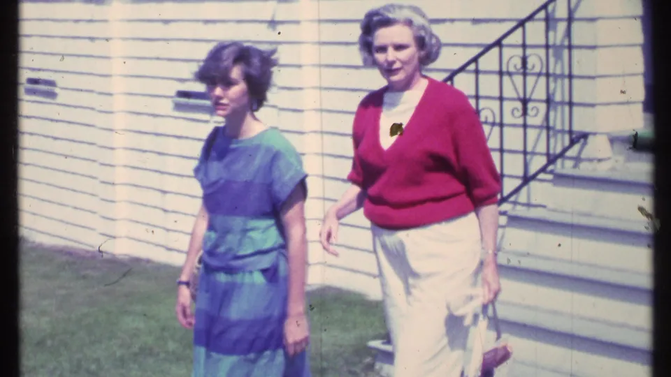 Two women walk together across a Vermont lawn in 1984, passing the front steps of a white clapboard house.