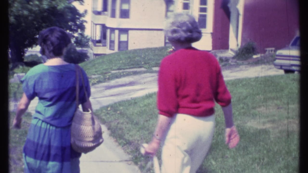 Two women walk away on a sidewalk beside grass and residential buildings.