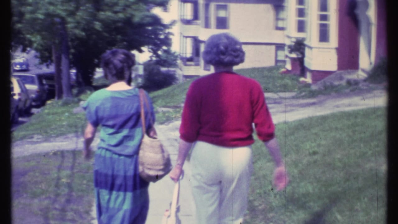 Two women continue down the sidewalk with parked cars and trees nearby.