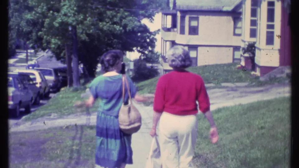 The women move farther down the suburban sidewalk, framed by cars, mature trees, grass, and nearby houses.