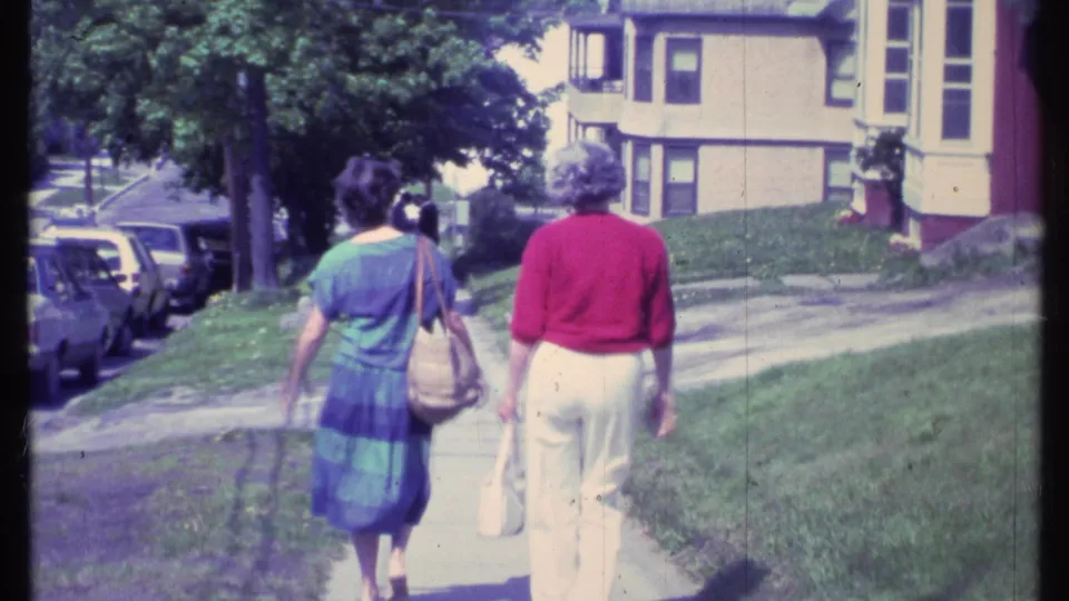 At the end of the clip, the two women continue away on the Vermont sidewalk with grass, trees, cars, and houses around them.