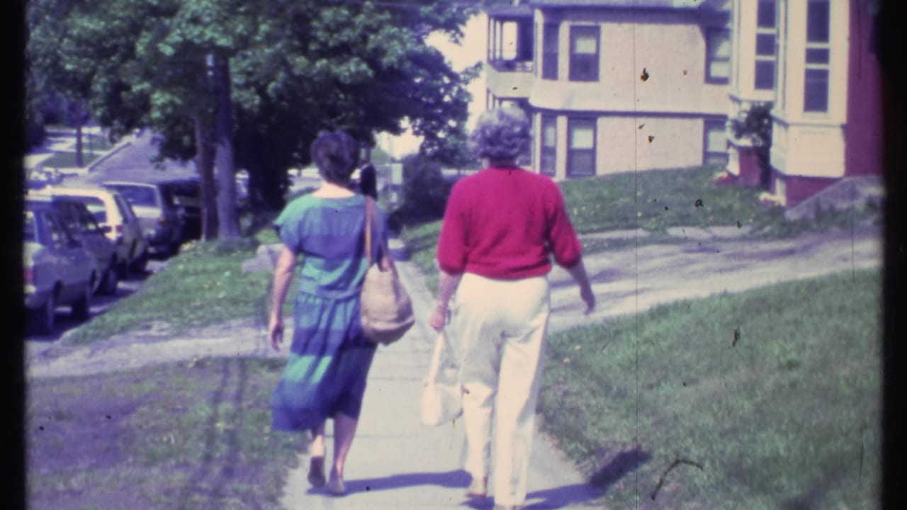 Two women walk away down a sunny sidewalk past parked cars and houses.
