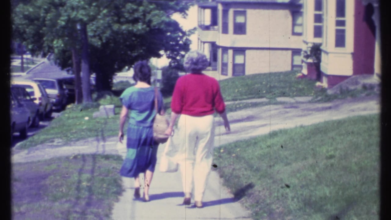 Two women continue walking away along a residential sidewalk beside grass and parked cars.